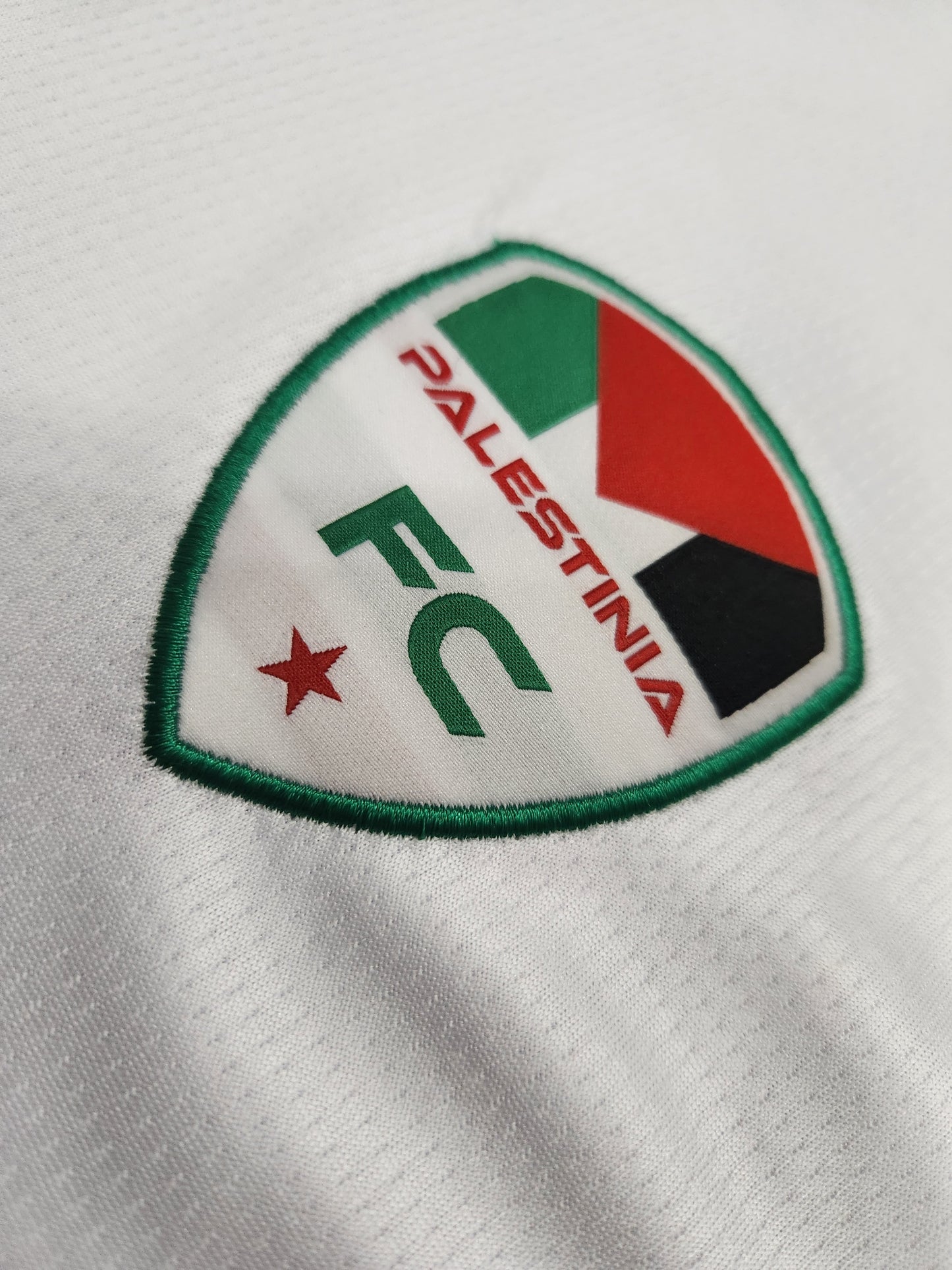 Close-up of Palestinia FC embroidered crest on white football jersey featuring Palestinian flag colors