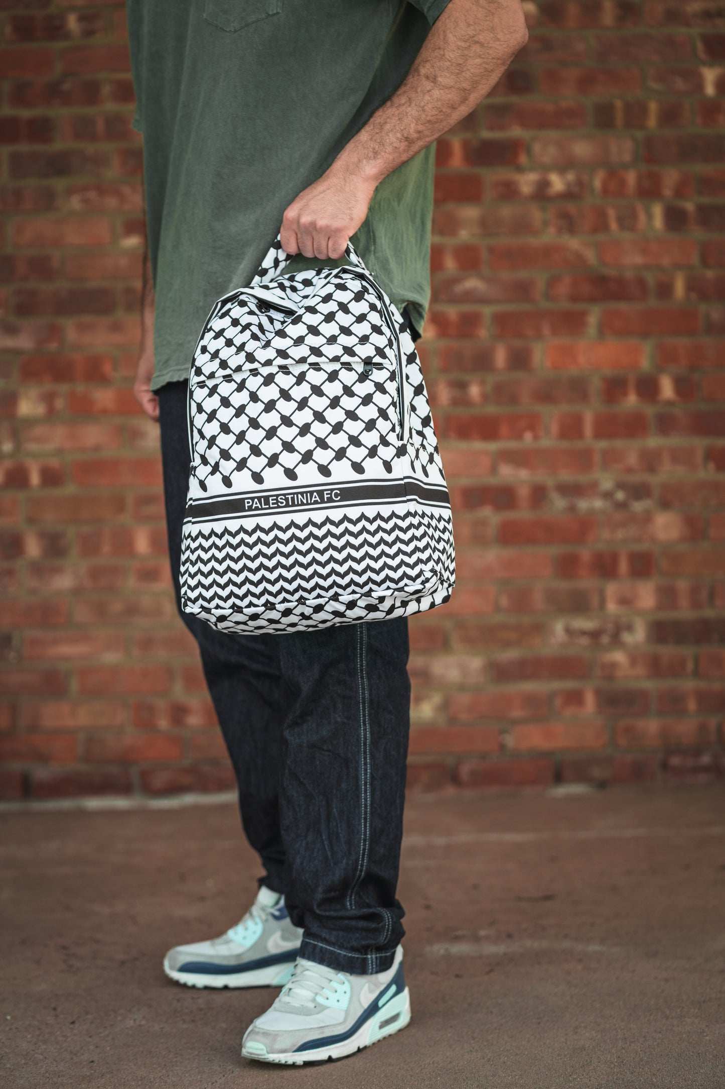 Person holding Palestinia FC Keffiyeh Backpack with kufiya pattern, symbolizing heritage and pride against brick wall background