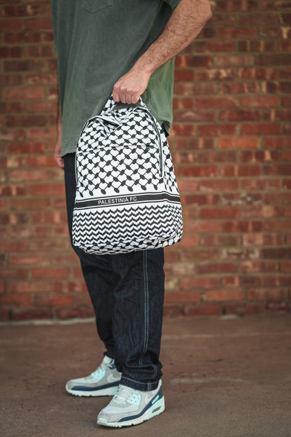 Person holding Palestinia FC Keffiyeh Backpack with kufiya pattern, symbolizing heritage and pride against brick wall background