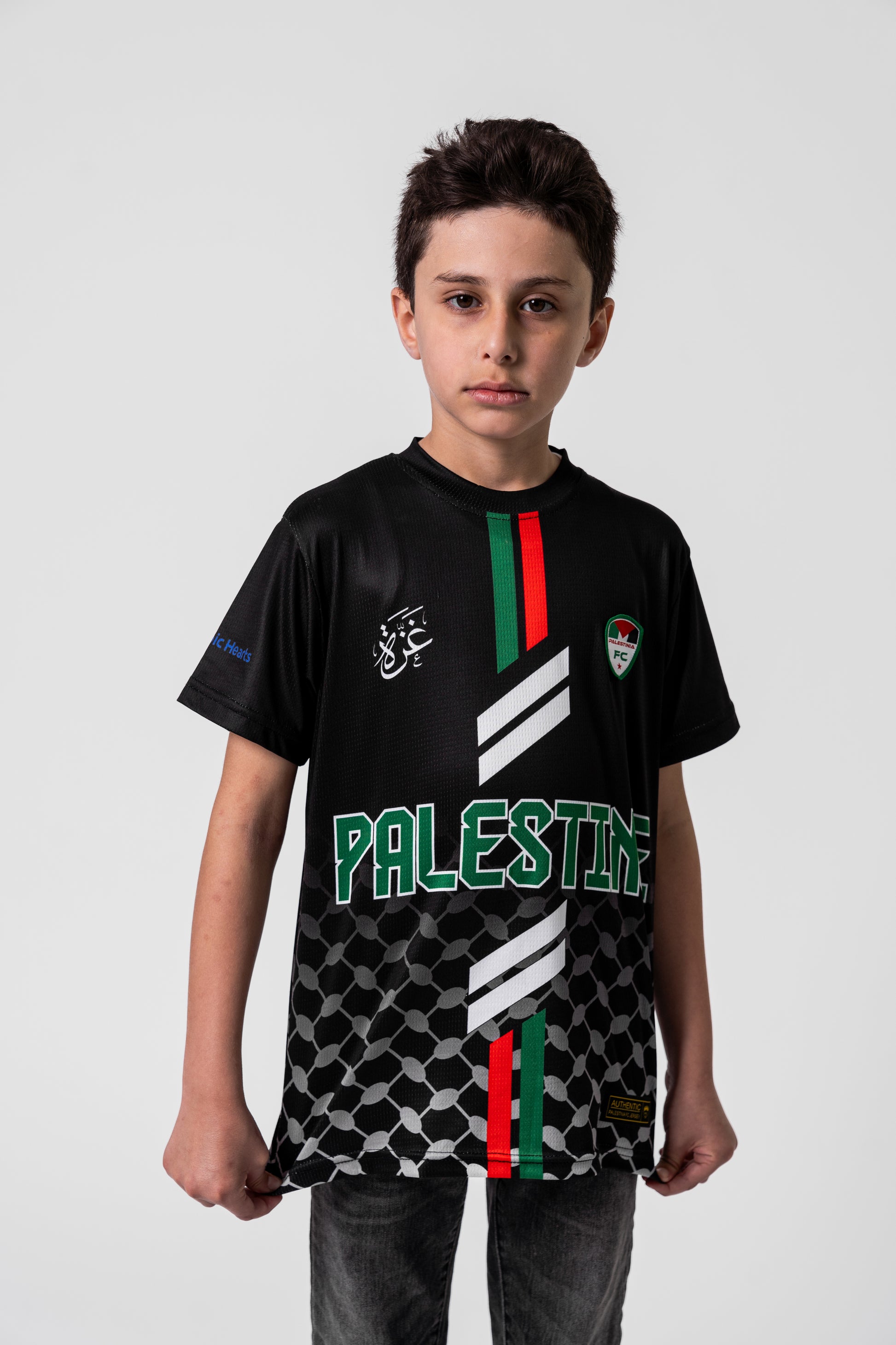 Boy wearing Kids Black Palestine football jersey with red and green stripes, keffiyeh pattern, and Palestinia FC crest