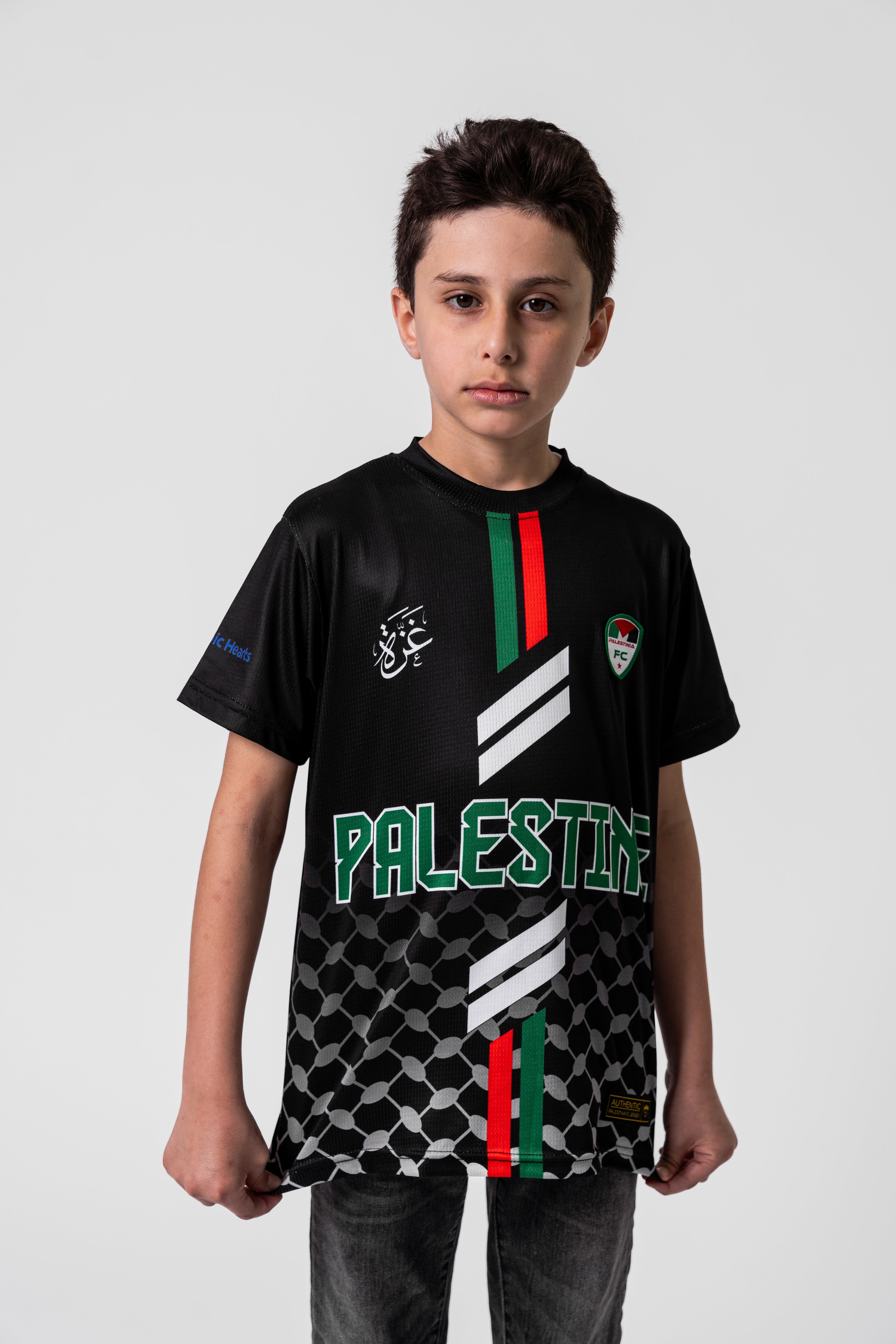 Boy wearing Kids Black Palestine football jersey with red and green stripes, keffiyeh pattern, and Palestinia FC crest