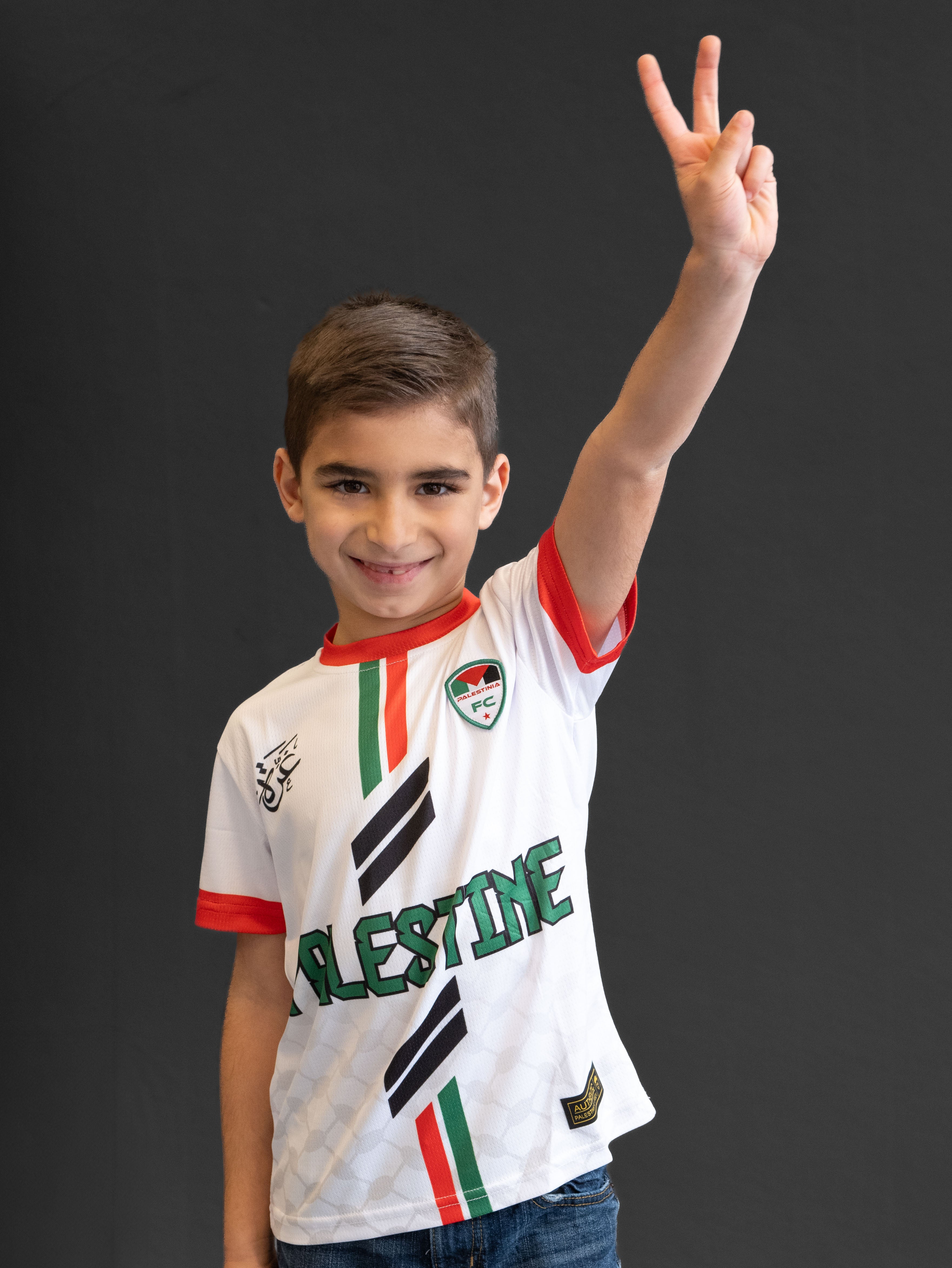 Smiling child wearing Kids White Palestinia FC jersey with red and green stripes, raising peace sign, symbolizing pride and unity