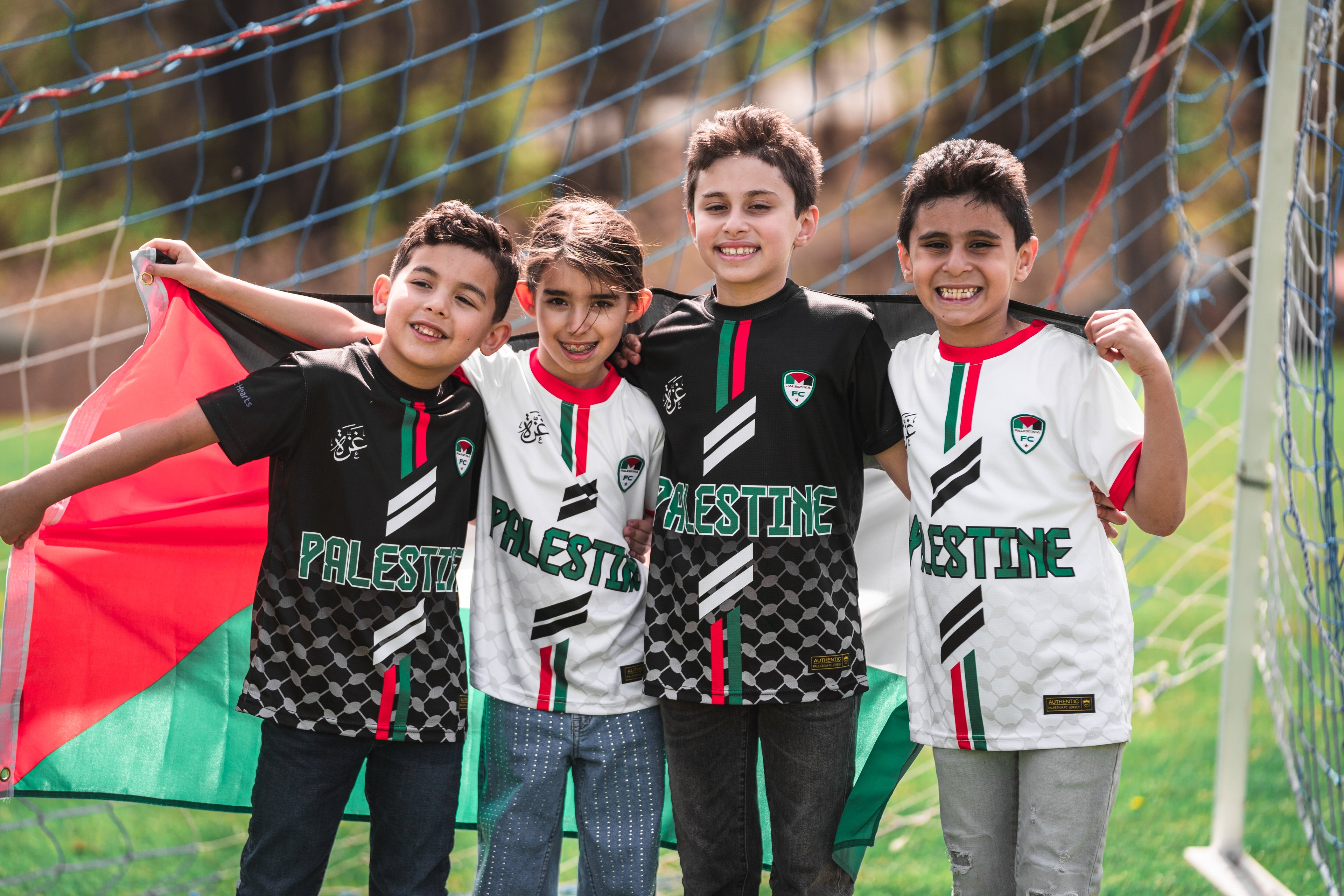 Four kids wearing Palestinian jerseys with a flag in the background on a soccer field.