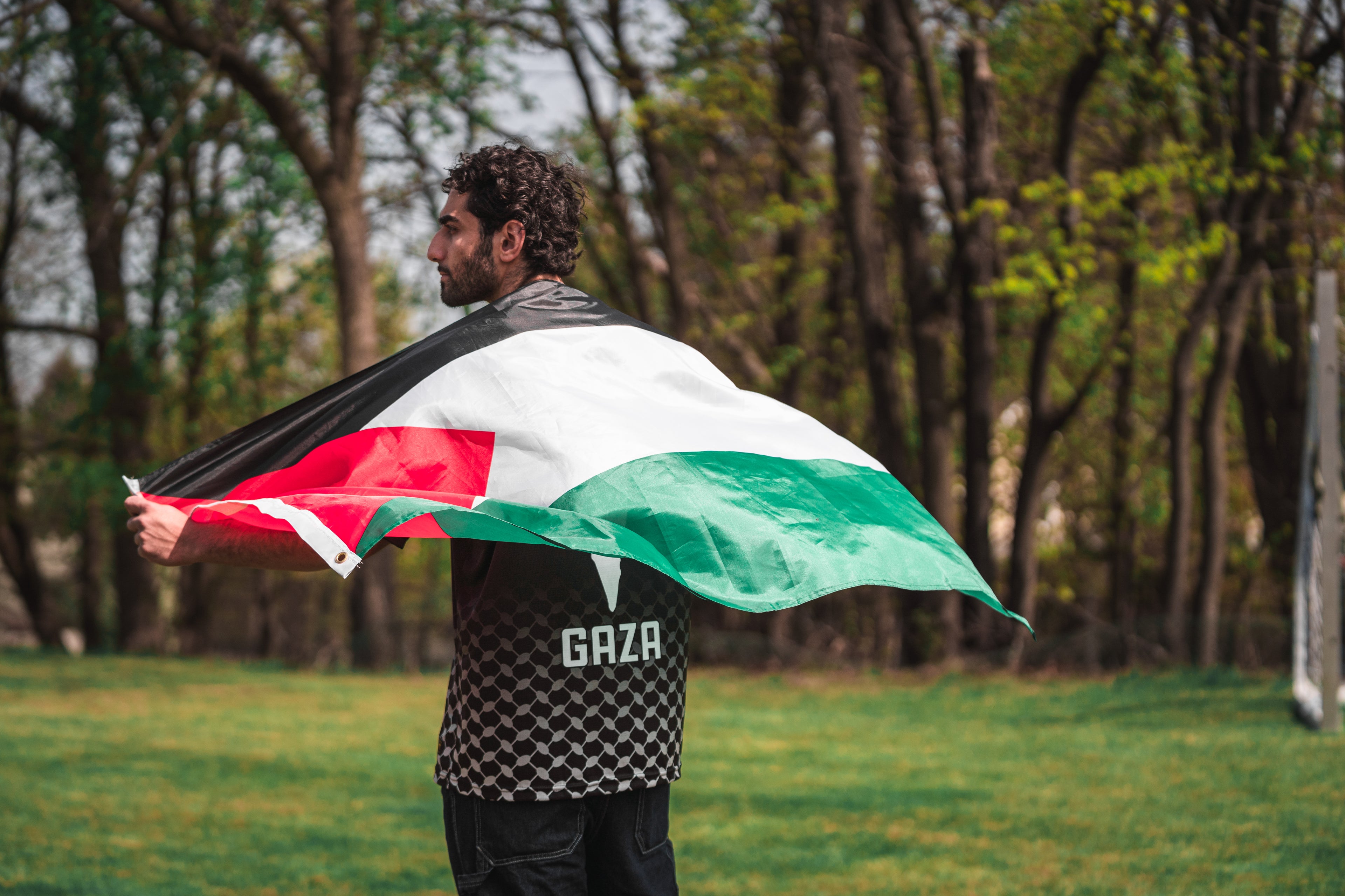 Person holding a Palestinian flag with a patterned shirt that says "Gaza" on the bottom in an outdoor setting